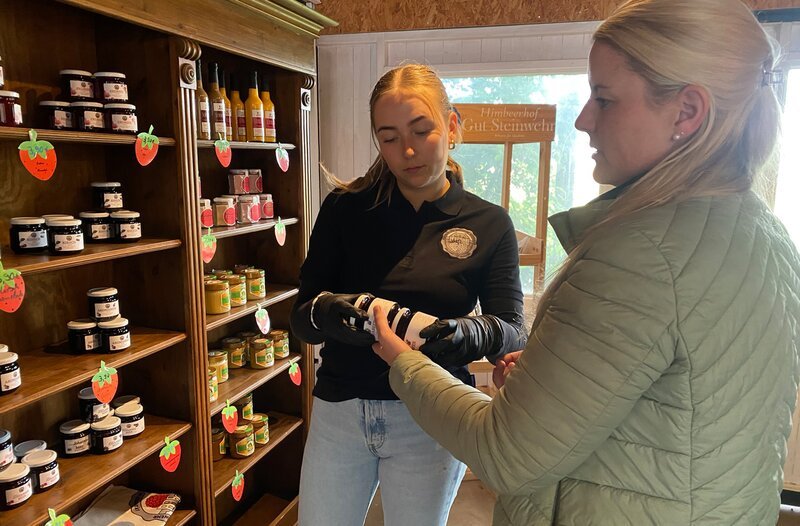 Judith Wannagat (rechts) vom Gut Steinwehr klärt im Hofladen Mitarbeiterin Hanna über die unbekannte Aronia-Beere auf, aus der Gelee gemacht wird. – Bild: NDR/​ Mario Göhring/​ Joker Pictures GmbH