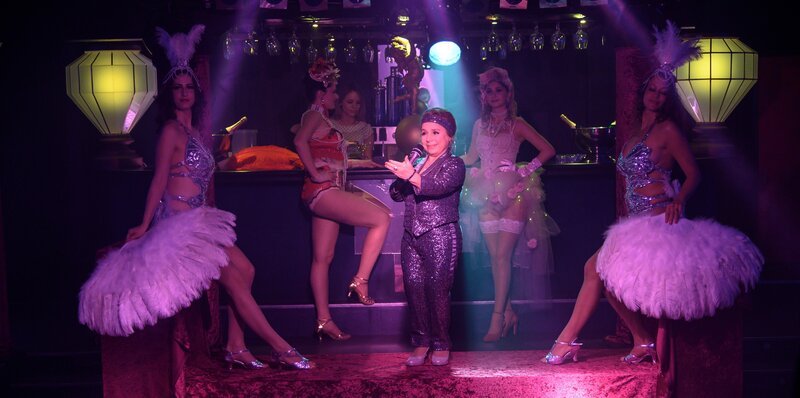 Verführung im „Salon Rouge“: Dora Krumme (ChrisTine Urspruch, M.), die Managerin des Burlesque-Theaters, moderiert ihre attraktiven Tänzerinnen (Komparsinnen) an – bald ist hier jedoch keinem mehr zum Tanzen zu Mute. – Bild: ZDF und MARKUS_FENCHEL
