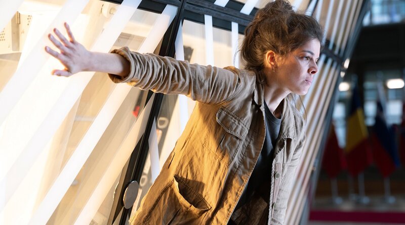 Die Umweltaktivistin Lydia (Anais Parello) hat sich während des EU-Gipfels an die Wand in der Lobby des Europäischen Rats geklebt. – Bild: WDR/​Cinétévé/​Studio Hamburg Serienwerft