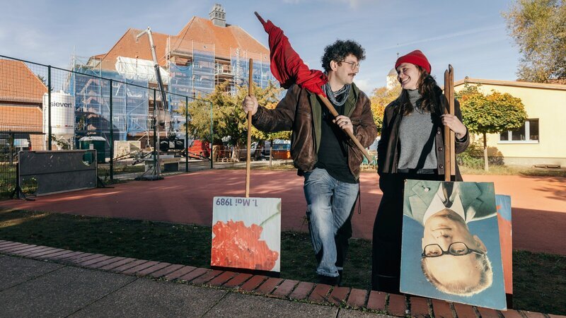 Bei der Sanierung entdecken Johannes und Louise noch alte Schätze aus der Zeit der ehemaligen DDR – Fahnen, Plakate und Bilder. Im Hintergrund ist das eingerüstete Grundschulgebäude zu sehen. – Bild: ZDF