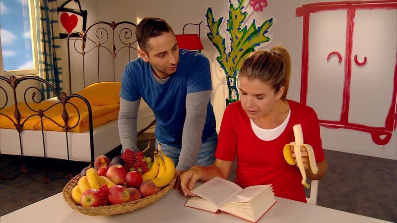Denis Moschitto würde sich so gerne eine leckere Banane von Anke Engelkes Obstteller nehmen, doch dafür muss er zuerst das Zauberwort nennen. – Bild: WDR/​Trickstudio Lutterbeck
