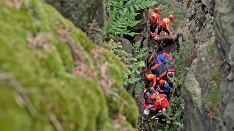 Eine Wanderin stürzt aus acht Metern Höhe in eine enge Felsspalte. Sie überlebt, ist aber verletzt. Das Sebnitzer Bergwacht-Team will sie retten, doch die Felsen stellen große Hindernisse dar. Jetzt sind die Künste und das Können der Felsretter gefragt. Die größte Herausforderung: die Patientin mithilfe von Seiltechnik und Muskelkraft so schonend und schnell wie möglich zwischen den Felsen heraus zum Rettungswagen zu bringen. – Bild: MDR/​Mia Media