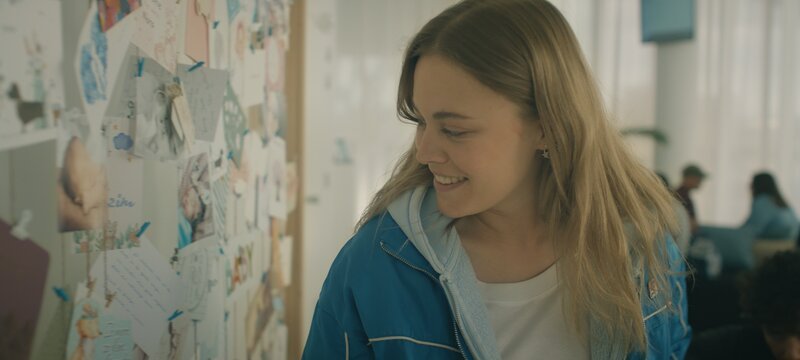 Charlie (Roos Dickmann) amüsiert sich im Wartebereich der Kinderwunschklinik über die ungewöhnlichen Namen einiger Kinder, die auf Geburtskarten zu lesen sind. – Bild: VRT, Les Gens and Gardner & Do /​ ZDF