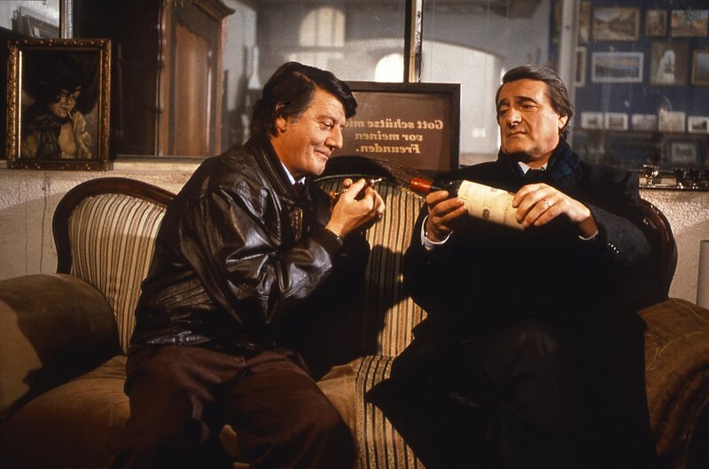 Im Auktionshaus probieren Josef Haslbeck (Helmut Fischer, rechts) und Schorschi Gruber (Hans Brenner) aus einer alten, wertvollen Weinflasche, die sie mit billigem Lambrusco wieder auffüllen wollen – doch beim ersten Schluck wird den beiden klar: irgendjemand ist ihnen zuvorgekommen. – Bild: BR/​Tatiana Film GmbH & Co. Audio KG