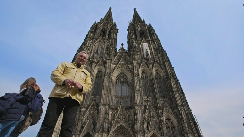 Wladimir Kaminer vor dem Kölner Dom: Im Vordergrund präsent, während die detailreiche Fassade des Wahrzeichens den Hintergrund dominiert. – Bild: 3sat