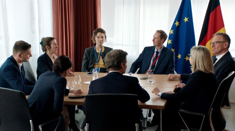 Die deutsche Delegation überlegt zwanghaft, wie sie den Vorwurf der anderen EU-Mitgliedsstaaten – Deutschland sei gespalten und handle ohne klare Linie – entkräften könnten. (Rosa Lembeck, Andreas C. Meyer) – Bild: WDR/​Cinétévé/​Studio Hamburg Serienwerft