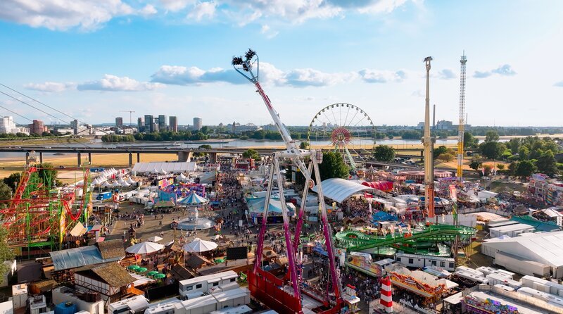 WDR Fernsehen LOKALZEIT LIVE – RHEINKIRMES 2025 IN DÜSSELDORF, am Freitag (17.07.26) um 20:15 Uhr. Die Düsseldorfer Rheinkirmes ist ein Ausnahmezustand auf Zeit: Wo eben noch eine leere Rheinwiese liegt, entsteht innerhalb weniger Wochen eine Stadt aus Stahl, Licht, Musik, Wohnwagen, Imbissbuden und tonnenschweren Fahrgeschäften. Der Film begleitet die „Größte Kirmes am Rhein“ von den ersten Pflöcken im Gras bis zum letzten Wochenende – und erzählt von den Menschen, die dieses Volksfest möglich machen. (Archivfoto: Luftaufnahme der Düsseldorfer Rheinkirmes 2025) – Bild: WDR/​Christian Hill/​erzaehlfabrik