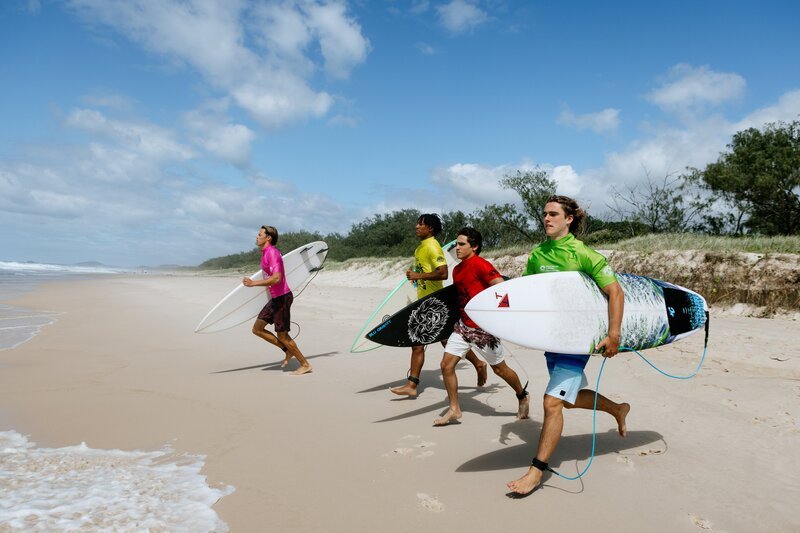 Griff (Mitchel Hardaker, linkls), Tommy (Marcel Laidlaw, 2. v. links), Marlon (Joao Marinho, 3. v. links) und Ari (Kai Lewins, rechts) rennen mit ihren Surfbrettern ins Wasser. Die nationalen Surfwettkämpfe haben begonnen und bisher läuft es für das Team eher schlecht. Das soll sich nun ändern. – Bild: ZDF/​Caitlin Miers
