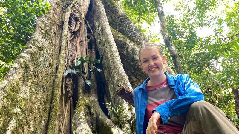 Nina am Fuß einer riesigen Würgefeige. Nachts tauchen an diesem Baum gern Wickelbären auf. – Bild: BR/​Text und Bild Medienproduktion GmbH & Co. KG/​Marcella Müller