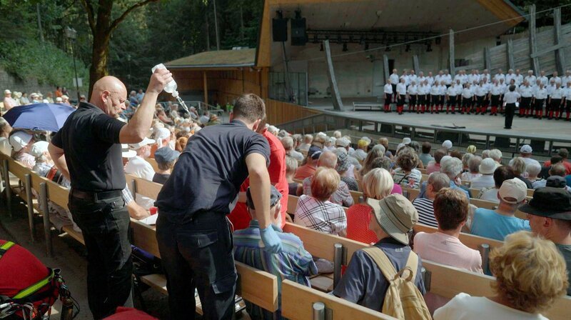 Der Sommer in der Sächsischen Schweiz ist heiß und trocken. Die Felsenbühne in Rathen ist dennoch gut besucht. Ein älterer Gast hat zu wenig getrunken und sein Kreislauf macht schlapp. Auch dafür sind die Bergretter da – denn sie sind mit ihrer Wache in Rathen am schnellsten da, wenn an der Freilichtbühne jemand in Not ist. Doch kaum sind die Retter am Patienten, kommt der nächste Alarm: ein zweiter Mann droht bewusstlos zu werden. Wassermangel der ganz anderen Art wenige Kilometer weiter rund um Bad Schandau: Rund um Schmilka brennt der Nationalpark großflächig. Bergretter Jost Bähne ist seit Tagen im kräftezehrenden Dauereinsatz. Als Fachberater lotst er die Feuerwehren durch den Nationalpark. Und koordiniert die Bergwacht, die bereitsteht, falls den Einsatzkräften etwas passieren sollte. – Bild: MDR/​Mia Media