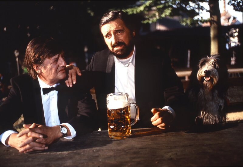Als Josef (Helmut Fischer, rechts) erfährt, wieviele Journalisten zur Eröffnung der Galerie „Haslbeck – Gruber“ kommen wollen, flüchtet er zum Unverständnis seines Kompagnon Schorschi (Hans Brenner) mit seinem Hund Lumpi in den Biergarten. – Bild: BR/​Tatiana Film GmbH & Co. Audio KG