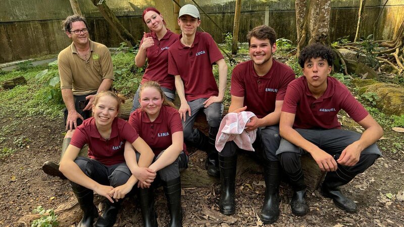 Michael Ernst, Anuk, Lina, Leonie, Luca, Emilio und Samuel (v. l. n. r.) sitzen auf Baumstamm nach der Otterbeckensäuberung im Zoo Leipzig. – Bild: MDR/​Cine Impuls Leipzig GmbH