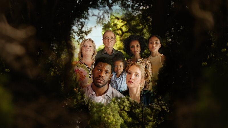 In einer grünen Hecke ist ein Loch, durch das man einen Blick hinter die Hecke werfen kann. Dort ist eine Gruppe von Menschen zu sehen: Ganz vorne die Köpfe von Dawit (Akeem van Flodrop, 2.v.l.) und Laura (Sina Martens, 3.v.r.). Hinter ihnen stehen Heidi (Gabriela Maria Schmeide, l.), Walter (Peter Lohmeyer, 3.v.l.), Fiyori (Lousia Heinricht, M.), Lemlem (Dennenesch Zoudé, 2.v.r.) und Armana (Chioma Chukwuani, r.). – Bild: Alina Simmelbauer /​ [M] NEUESUPE /​ ZDF