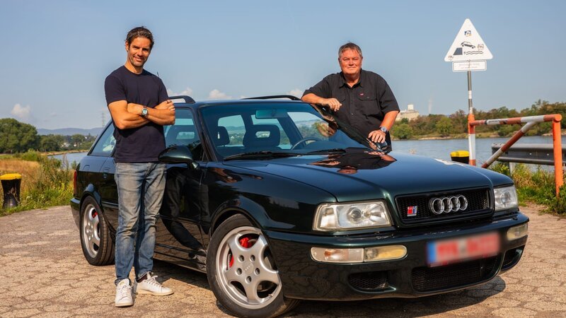 Mike standing on right of Audi RS2, Elvis standing on left of Audi RS2, both with arms resting on car smiling looking into stills camera,, lake in background – Bild: Warner Bros. Discovery