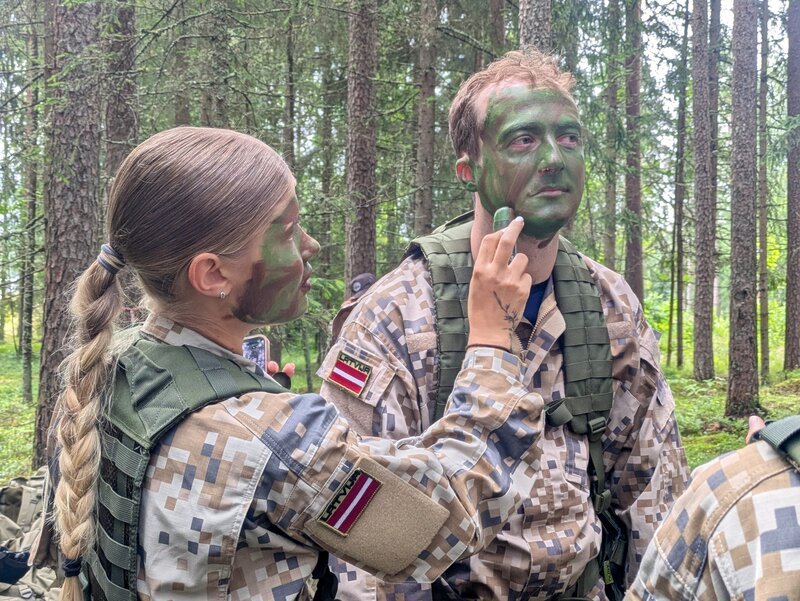 Während eines freiwilligen militärischen Trainings in einem Sommercamp für Schülerinnen und Schüler in einer Kaserne der lettischen Armee bei Alūksne (Marienburg) im Nordosten Lettlands trägt eine Teilnehmerin dem quer-Reporter Philipp Potthast (rechts) Tarnschminke auf. – Bild: BR/​Viola Nowak