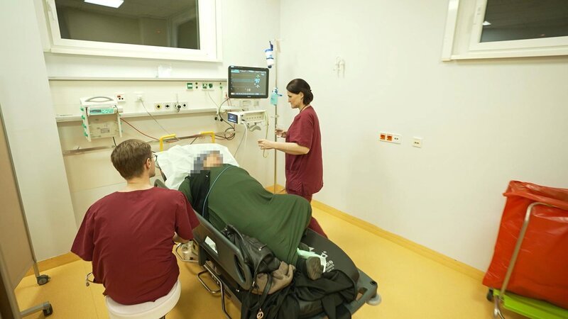 Berlin: two members of staff are around a laying patient. The male one is sitting on a white stool, the female one is standing at the head of the bed seemingly bringing the patient something to drink. To the right we see a big trash container with a red bag. – Bild: Warner Bros. Discovery /​ Production Company Staff