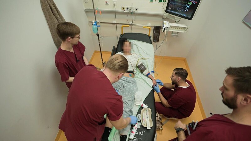 Berlin: three members of staff are standing, another one kneeling around a female patient. One of the nurses is at her feet, while the other is checking her hand probably prior to extract some blood. – Bild: Warner Bros. Discovery