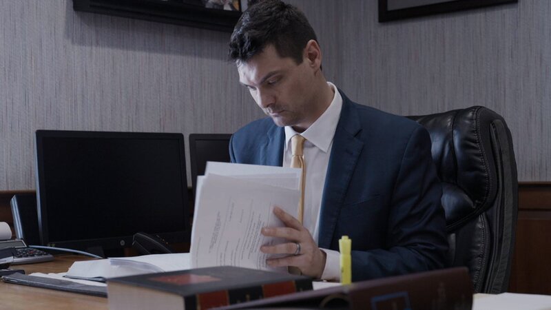 Contributor Clayton Crates, Judith Hawkey’s Defense Attorney, sat at this desk looking through paperwork, with a book and folder on the desk in the foreground. Interior, landscape – Bild: Warner Bros. Discovery, Inc. or its subsidiaries and affiliates
