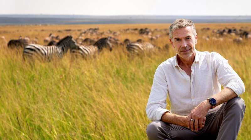 Dirk Steffens sitzt vor einer Zebraherde in der Masai Mara in Kenia. Millionen von Gnus und Zebras ziehen hier jedes Jahr durch die Savanne; eine der größten Landwanderungen von Tieren überhaupt. – Bild: NDR/​ARD/​Doclights/​Oliver Rötz