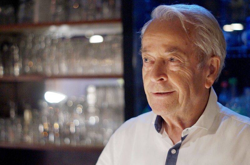 Ein alter Mann mit weißem Hemd steht in einer dunklen Gaststätte vor einem Gläserregal und lächelt. Achim Wenning ist 82 und immer noch Gastwirt mit Leib und Seele. – Bild: MDR/​Lucas Schwarz