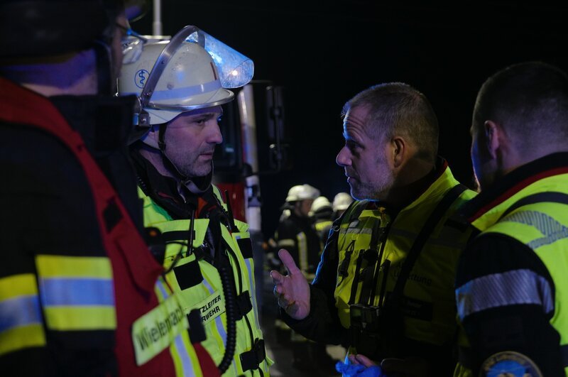 Dr. Bender (2.v.l.) bespricht sich mit einem Feuerwehrmann am Unfallort. – Bild: me:works /​ ZDF und me:works