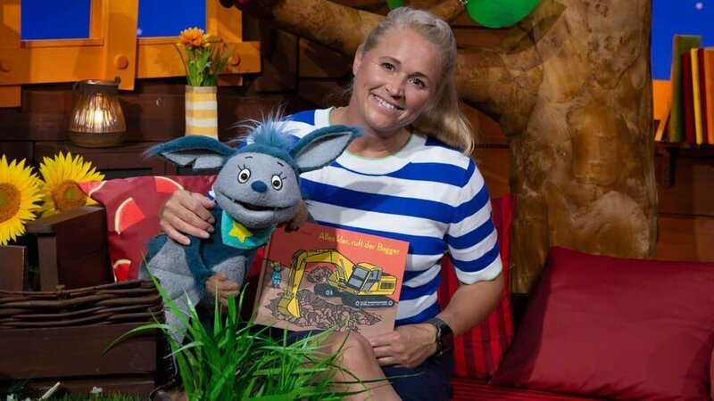 Singa stellt ein Buch aus der Vorlesezeit vor. Fidi wird neugierig: Was passiert auf einer Baustelle? – Bild: KiKA