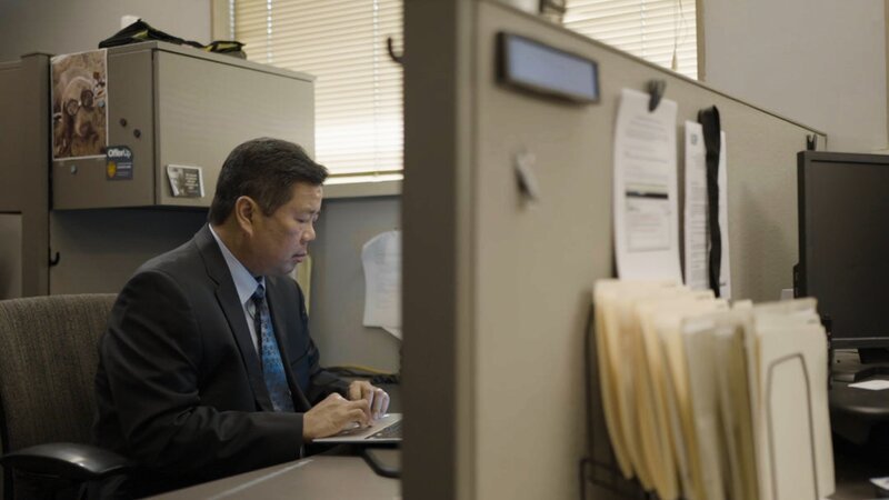 Contributor Thien Do, Lead investigator, sat at his desk looking at his laptop in front of him. There is a an office cubicle divider in front of him, with a computer, stack of paperwork and water bottle on the other side of it, out of focus. Interior, landscape – Bild: Warner Bros. Discovery, Inc. or its subsidiaries and affiliates