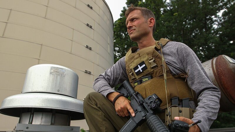 John kneeling while spying on Terry with assault rifle in his lap. – Bild: Discovery Channel (ROMA) /​ Colby Hunt