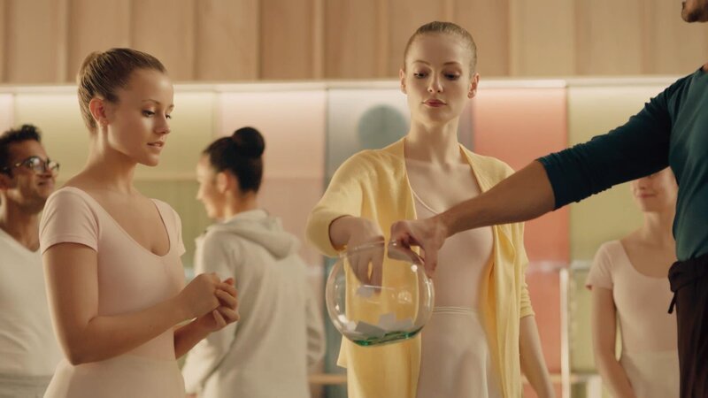 Für die heutige Aufgabe im Ballettunterricht sollen Paare gebildet werden. Jeder darf einen Zettel mit dem Namen des Tanzpartners ziehen. Thea (M. Hanna Dodd) fischt gerade einen Zettel aus dem Glas, das Lehrer Armando Castillo (Rik Young, r.) hält, während Lena (Jessica Lord, l.) sehr gespannt darauf wartet, an der Reihe zu sein. – Bild: ZDF/​Cottonwood – Opéra national de Paris – ZDF – ZDFE. Picture Thibault Grabherr
