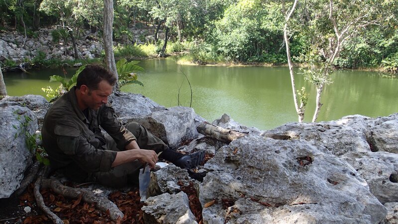 John Hudson sits on bank rocks in the shade of jungle trees while purifying water. – Bild: Animal Planet