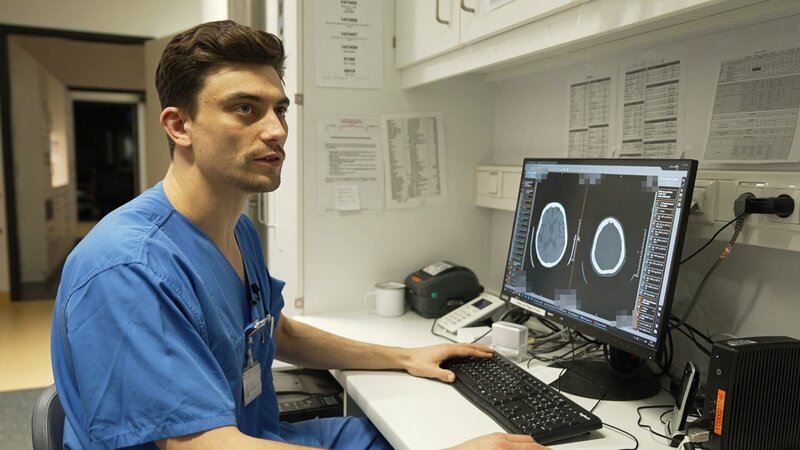 Rostock: young physician Max is sitting at a desk while giving an interview and explaining what he sees on the CT images of a skull on his computer screen. He is seemingly looking at the interviewer, while his hands are on the keyboard and the computer mouse. – Bild: Warner Bros. Discovery /​ Production Company Staff