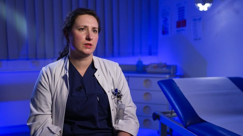 Lübeck: Assistant Surgeon Olga gibt ein Interview am Interview-Set. Sie trägt dunkelblaue OP-Kleidung und einen weißen Kittel; links von ihr befindet sich eine blaue, mit einem Papierlaken bedeckte Matratze. In ihrer Tasche sind unter anderem Scheren zu sehen. – Bild: Warner Bros. Discovery /​ Production Company Staff
