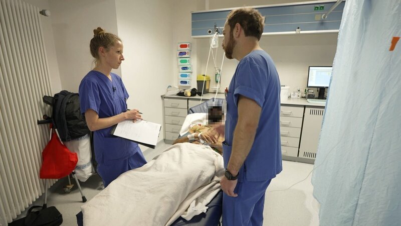 Saarbrücken: one young doctor and another male physician both in blue scrubs are standing around a pixelated laying patient. The young doctor is intently listening to what her colleague says. To the right we see blue curtains, and in the back some light grey cabinets. – Bild: Warner Bros. Discovery /​ Production Company Staff