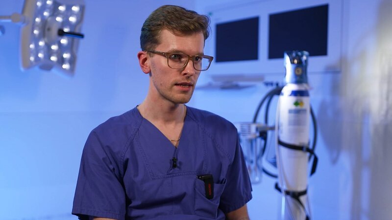Mönchengladbach: Der junge Arzt Lorenz beantwortet während eines Interviews scheinbar eine Frage. Hinter ihm befinden sich eine große OP-Leuchte und eine Sauerstoffflasche. Das Licht im ITV-Set ist bläulich. – Bild: Warner Bros. Discovery /​ Production Company Staff