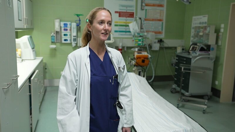 Lübeck: Neurologin Valentina gibt scheinbar ein Interview. Sie trägt einen weißen Kittel über blauen OP-Kleidern und befindet sich im Schockraum. Im Hintergrund ist ein leeres Bett mit weißem Laken zu sehen; der Raum ist grün gefliest. Links stehen hohe Schränke, im Hintergrund unter anderem medizinische Geräte und ein Rollwagen. – Bild: Warner Bros. Discovery /​ Production Company Staff