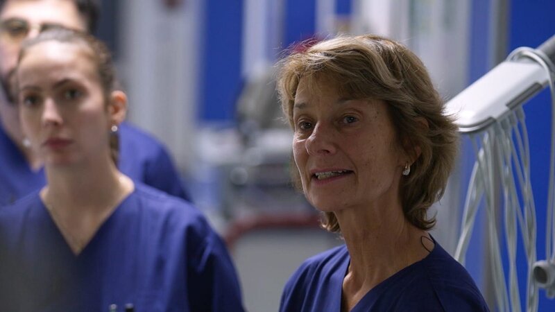 Düsseldorf: close-up shot Dr. Koenig is seemingly talking to the patient who has just arrived. In her scrubs pocket we see several ball pens and her green sight glasses. To her right her team is listening intently. Behind her we partially what seems to be an ECG device from which cables are hanging. – Bild: Warner Bros. Discovery /​ Production Company Staff