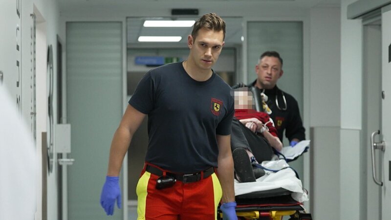 Herne: dynamic shot of two paramedics moving a gurney on the aisle. The one in the front is wearing a black T-shirt, red-yellow trousers and blue gloves and looking to the side. On the left we see the back of a physician in his white coat. – Bild: Warner Bros. Discovery /​ Production Company Staff