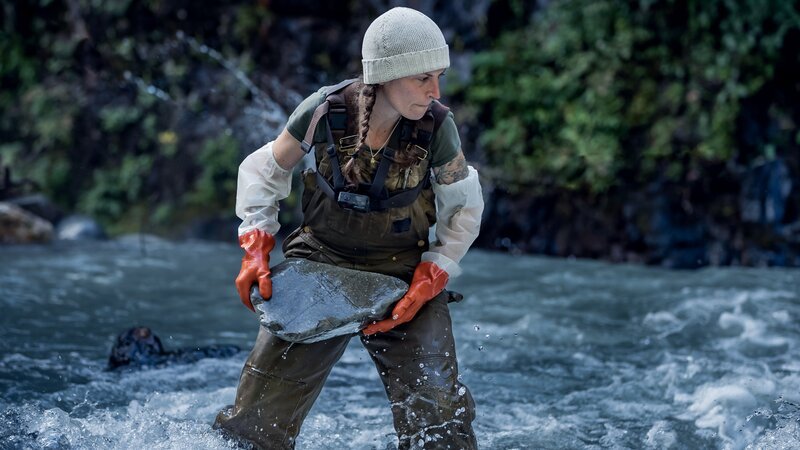 Kayla Johanson mit Schutzhandschuhen beim Steinwurf im Bach – Bild: Discovery, Inc. All Rights Reserved.