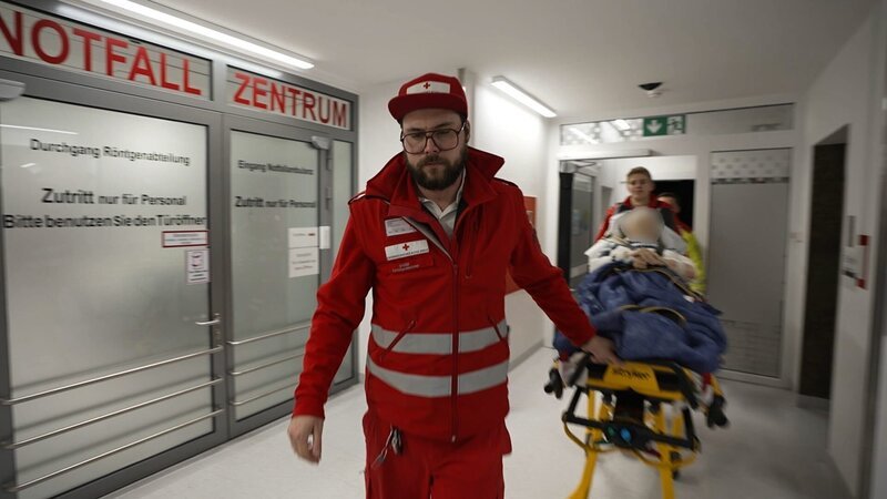 Füssen: dynamic shot of a bearded paramedic wearing all red, a cap and glasses pulling a gurney with a new pixelated patient into the A&E. To his right we see in big red letters the word for A&E Center in German over glass doors. Behind him and somewhat blurry we see a laying patient and a colleague of him. – Bild: Warner Bros. Discovery /​ Production Company Staff