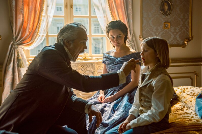 Kronprinz Rudolf (Arian Wegener, r.) wird im Beisein seiner Mutter Sisi (Dominique Devenport, M.) von Dr. Griebe (Vytautas Kaniuonis, l.) untersucht. – Bild: ORF/​Beta Film/​RTL/​Story House Pictures/​Armands Virbulis