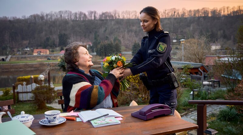 Janas Oma Babicka (Swetlana Schönfeld, l.) wurde Opfer eines perfiden Enkeltrickbetrugs. Jana (Barbara Prakopenka, r.) bringt ihr einen Blumenstrauß und fragt, wie es ihr nach dem Schock geht. – Bild: ARD/​Rudolf Wernicke