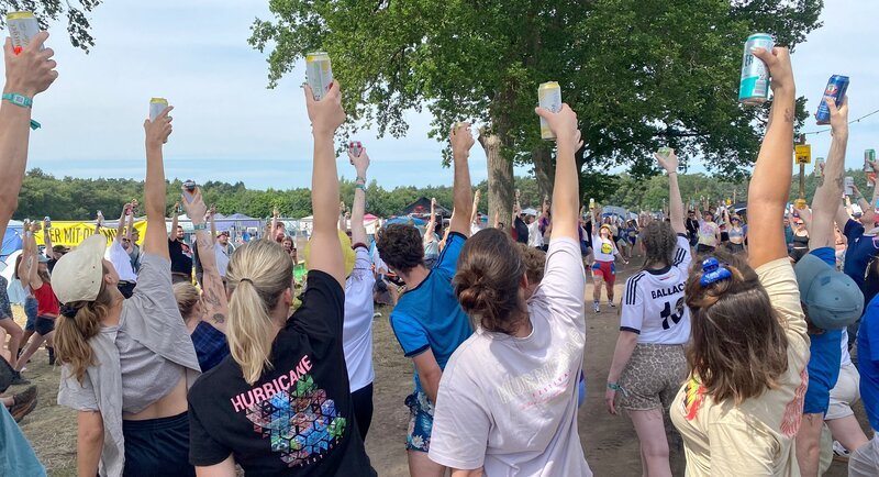 Bier-Yoga auf dem Campingplatz. Lynn macht die müden Festivalbesucher jeden Morgen um 11:00 Uhr mit Bier-Yoga fit für den neuen Festival-Tag. Dabei gibt es immer wieder einen erfrischenden Schluck. – Bild: NDR/​Filmteam Papstein