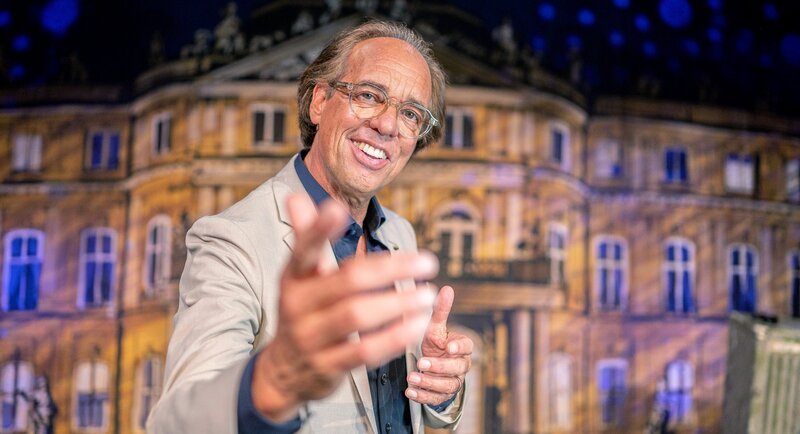 Christoph Sonntag steht grinsend auf der Bühne des Stuttgarter Wilhelma-Theaters und zeigt mit dem Finger in Richtung der Kamera. Im dritten Teil seines neuen Programms spricht er über Glück, Musik und Erinnerungen an die gute, alte Zeit. – Bild: SWR/​Christian Tichy