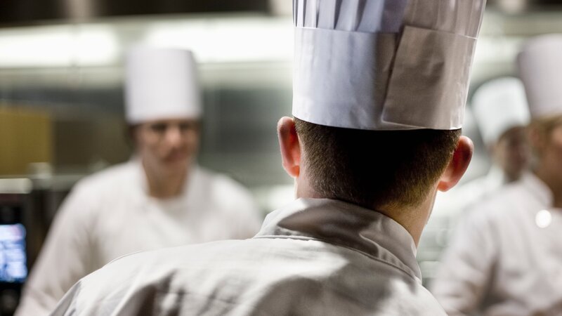A closeup view from behind of a chef wearing a toque hat in a commercial kitchen. – Bild: Mint Images /​ Getty Images