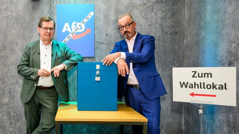 Ralf Stegner (l.) und Jan Fleischhauer (r.) im Wahllokal-Setting – zwischen blauer Wahlurne und einem Wegweiser zum Wahllokal. – Bild: ZDF
