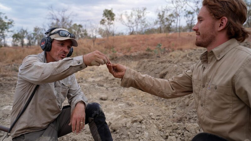 Andreas Macherey reicht seinem Sohn Tobi ein Goldnugget, das er gerade auf einem Goldfeld im australischen Northern Territory gefunden hat. – Bild: Warner Bros. Discovery /​ Thomas Danebrock