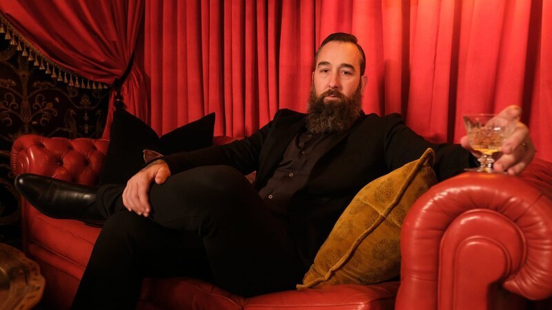 Homeowner Arryn holds a mixed cocktails while posing in his former embalming room, now a converted speakeasy, as seen on HGTV’s We Bought a Funeral Home. – Bild: Warner Bros. Discovery /​ Darren Goldstein