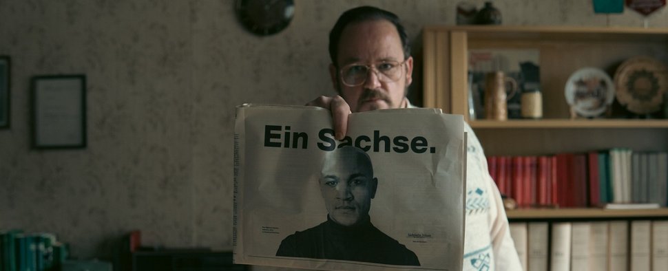 Schmidt (Andreas Enke) mit einem Zeitungsartikel über Samuel (Malick Bauer) in „Sam – Ein Sachse“ – Bild: Yohana Papa Onyango/UFA Fiction